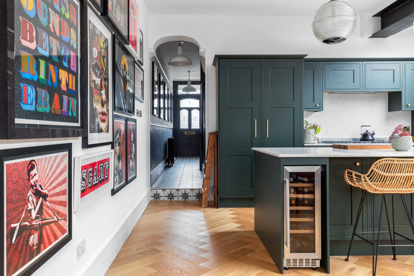 Kitchen with gallery wall of prints leading to hallway, wine fridge and rattan bar stool — interior photography for Architecture:WK, London