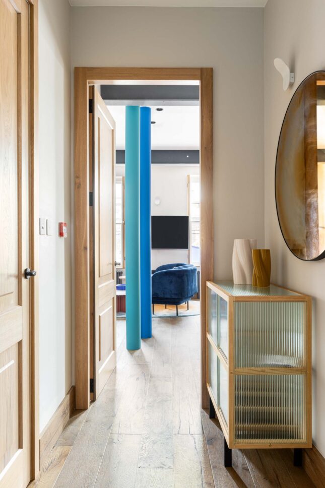 Office hallway with glass cabinet and round mirror leading to lounge, designed by Unknown Makers — Kingsland project, London